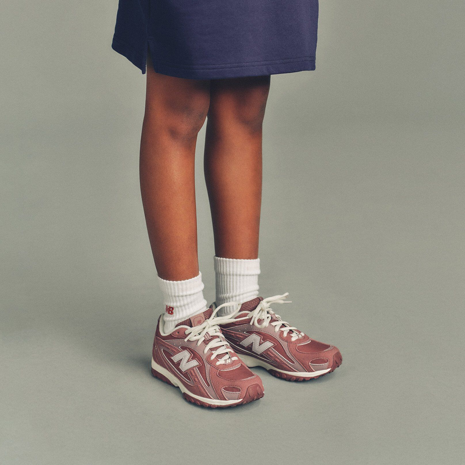 The child is wearing a navy top that extends to mid-thigh and white ribbed crew socks featuring a small red &ldquo;NB&rdquo; logo near the cuff