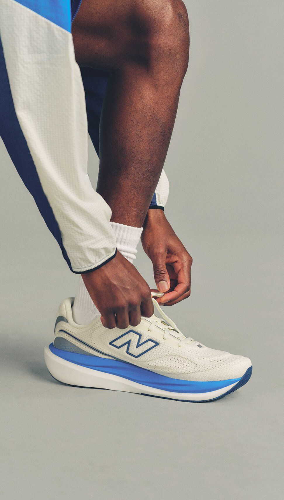 Close-up of a person tying white New Balance running shoes with blue accents while wearing white socks and light-colored athletic pants.