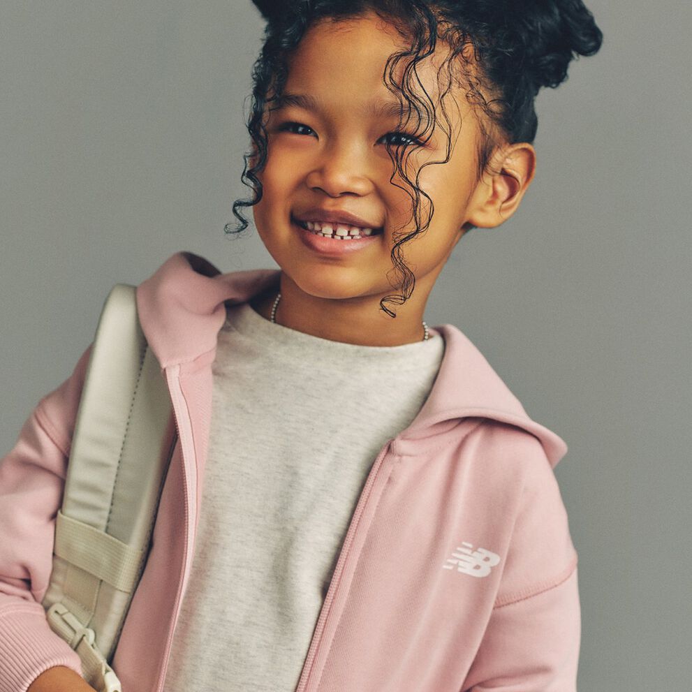The child is wearing a light gray T-shirt layered under a soft pink zip-up hoodie with a white New Balance logo on the chest.