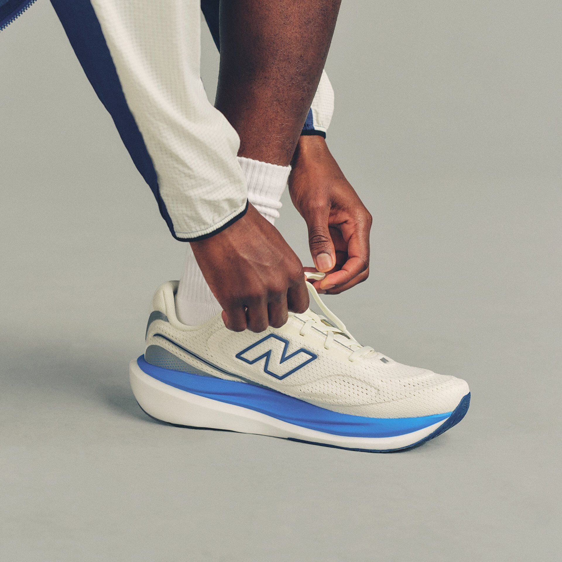 Close-up studio image of a person bending down to tie the laces of a white New Balance running shoe against a plain light gray background.