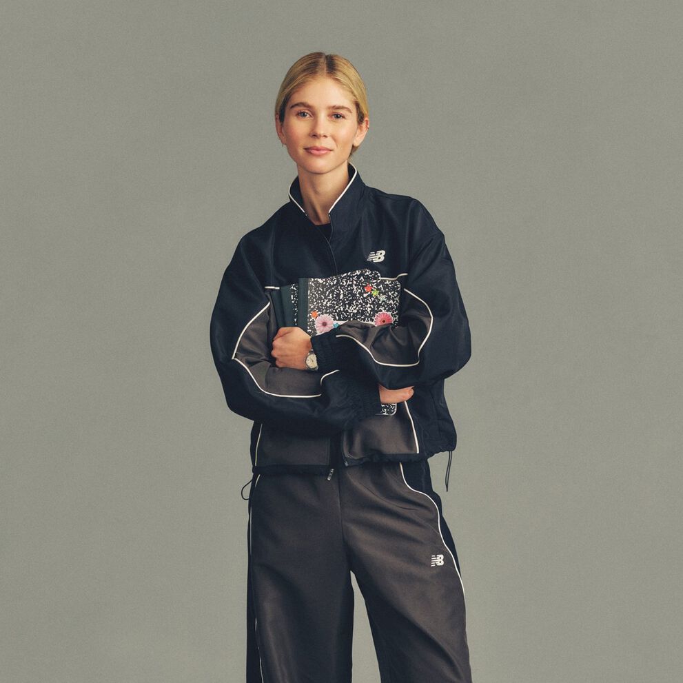 Studio image of a single person standing against a plain light gray background, wearing a dark track jacket and matching track pants with white piping and small &ldquo;NB&rdquo; (New Balance) logos on the chest and thigh.