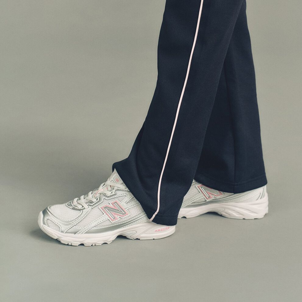 The pants fall loosely over a pair of white New Balance sneakers featuring mesh panels, layered overlays, and a large &ldquo;N&rdquo; logo outlined in soft pink on the sides. 
