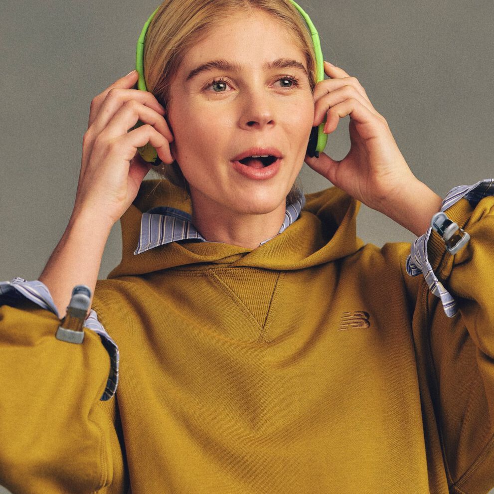 Close-up studio image of a person wearing a mustard-yellow hoodie with a small New Balance logo on the chest, layered over a striped collared shirt.