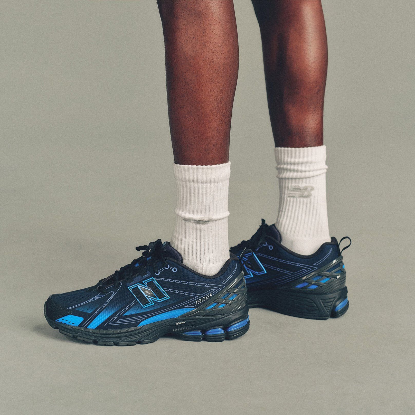 The person is wearing white ribbed crew socks and black New Balance running shoes with bright blue accents and a large outlined &ldquo;N&rdquo; logo on the sides. 