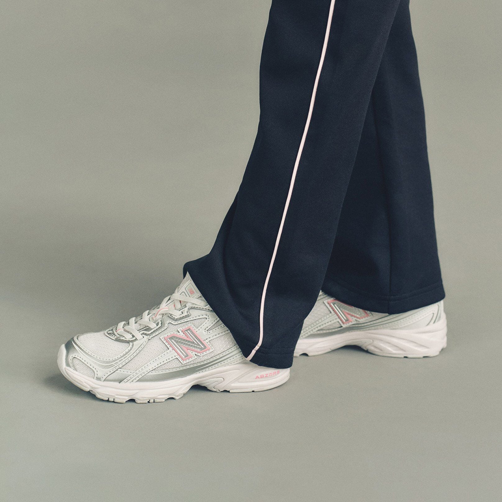 The pants fall loosely over a pair of white New Balance sneakers featuring mesh panels, layered overlays, and a large &ldquo;N&rdquo; logo outlined in soft pink on the sides. 