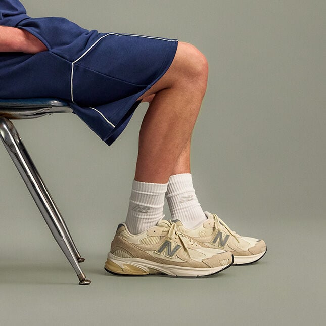 Studio image of a person seated on a metal chair, shown from mid-thigh down against a smooth, light gray background. 